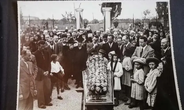 Una folla partecipa a un funerale, con un feretro fiorito al centro. Questa foto post mortem cattura un momento di lutto collettivo.