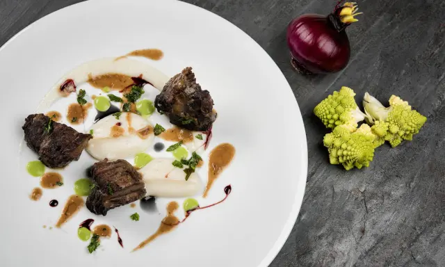Succulenta guancia di manzo tenerissima, servita su purè bianco con gocce verdi e salsa bruna. Accanto, cipolla rossa e cavolfiore romanesco.