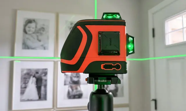 Pomarańczowa poziomica laserowa z zielonymi liniami na statywie, na tle oprawionych zdjęć.