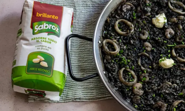 Arroz negro con calamares y gambas en paellera, junto a un paquete de arroz redondo Sabroz.