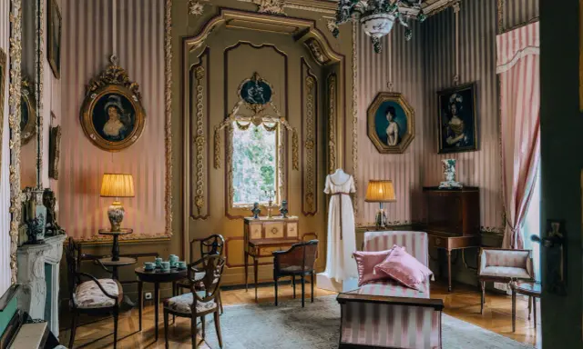 Eleganckie wnętrze pałacu w Nieborowie. W pokoju z różowo-białymi pasami na ścianach, zabytkowe meble, obrazy i manekin w sukni.