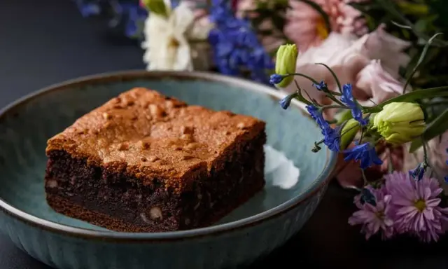 Un délicieux brownie healthy, parsemé de noix, dans un bol bleu, avec un bouquet de fleurs colorées en arrière-plan.