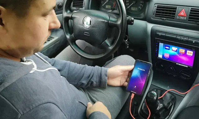Mężczyzna w samochodzie pokazuje, jak połączyć telefon z autem za pomocą kabla i aplikacji Weblink. Na ekranie radia widoczne są ikony aplikacji.