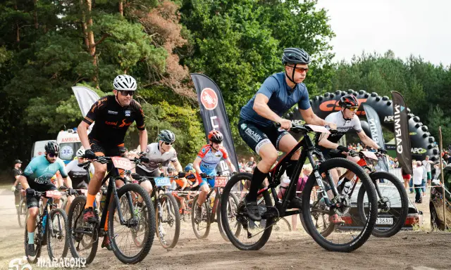 Kolarze biorą udział w wyścigu **zawody MTB**. Widać ich w akcji, jadących po piaszczystej trasie, z lasem w tle.