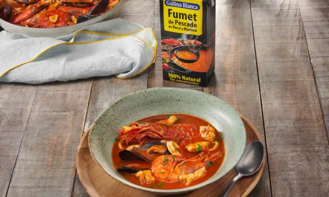 Un plato humeante de zarzuela de pescado, con gambas, mejillones y calamares, junto a un cartón de fumet de pescado.