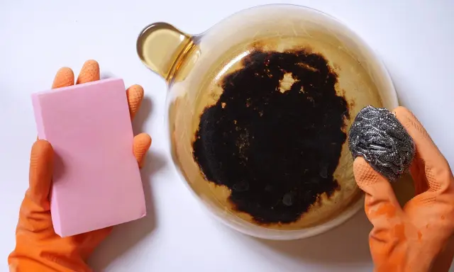 Manos con guantes naranjas sostienen una esponja rosa y un estropajo metálico, listos para aprender como quitar el quemado de una olla.