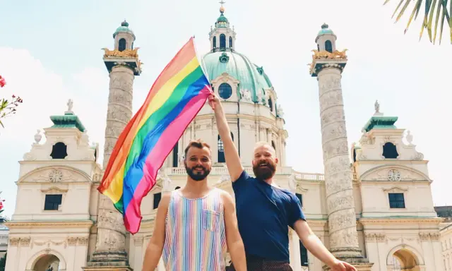 Two men proudly hold a rainbow flag in front of a grand building in Austria, celebrating gay pride.