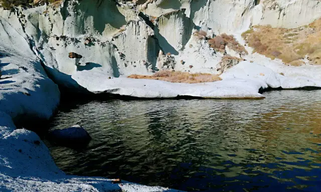 Scogliere bianche erose dal mare, con vegetazione secca e un'acqua scura che riflette il cielo. Sembra un luogo selvaggio, quasi un "cane malu" che custodisce la costa.