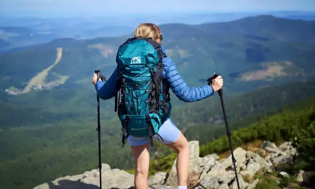 Kobieta z turkusowym plecakiem w góry, z kijkami trekkingowymi, podziwia panoramę.