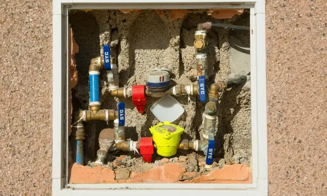 Instalacja wodna z pionowym montażem wodomierza, zaworami i rurami w ścianie.
