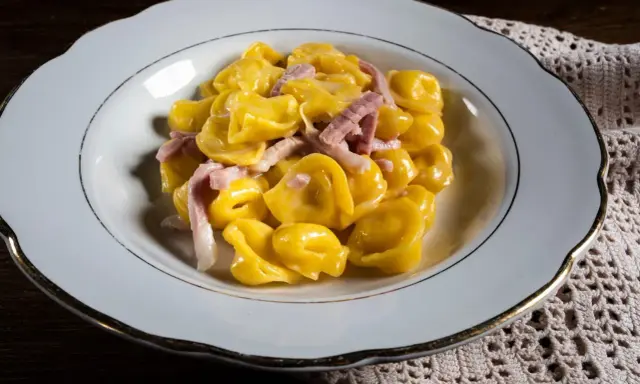 Tortelli ripieni in salsa cremosa con prosciutto, un classico tra i piatti tipici di Parma, serviti in un piatto bianco bordato d'oro.