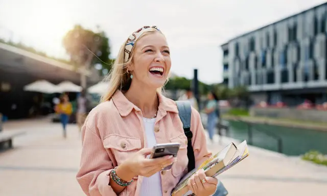 Szczęśliwa studentka z polską maturą, która planuje studia za granicą, śmieje się, trzymając telefon i książki.