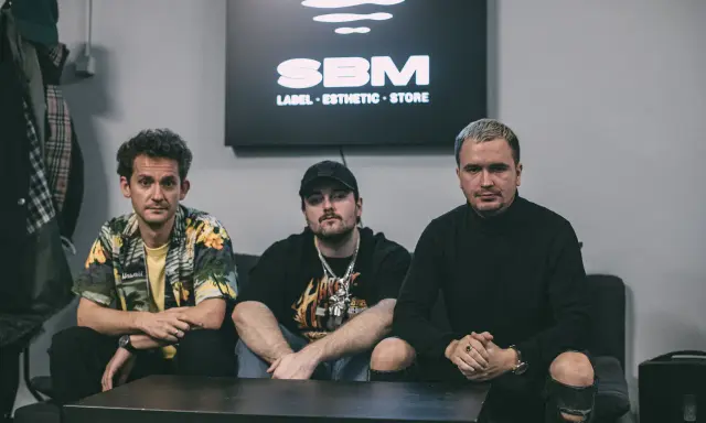 Trzech mężczyzn siedzi na kanapie. W tle logo SBM. Może to scena z teledysku lub sesji nagraniowej piosenki Bedoesa.