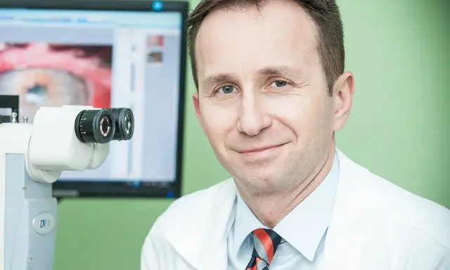Okulista z uśmiechem przy aparaturze do badań. Na ekranie widok oka, sugerujący diagnostykę przed przeszczepem rogowki.