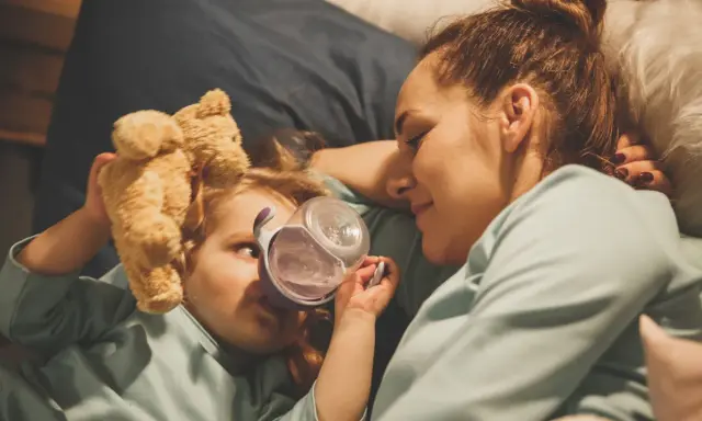 Mama opiekuje się dzieckiem z rotawirusem. Podaje mu wodę z kubeczka, a obok leży pluszowy miś.