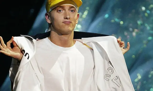 Slim Shady w żółtej czapce i białej bluzie, z gestem dłoni.