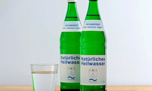 Zwei grüne Flaschen Heilwasser und ein Glas Wasser. Ein Glas Wasser vor dem Schlafengehen trinken kann beim Abnehmen helfen.