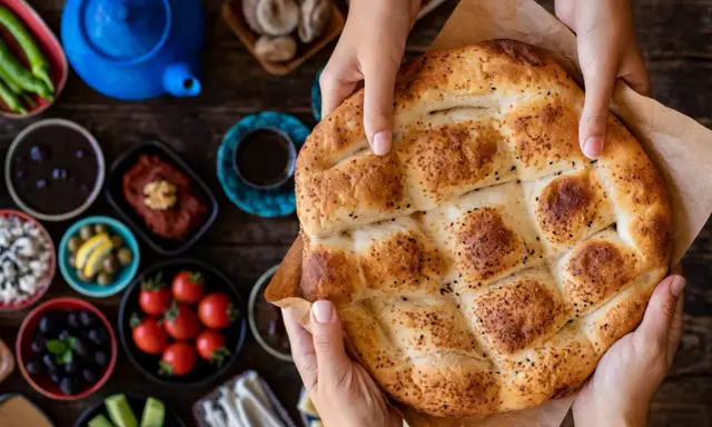 Hände halten ein frisch gebackenes Ramadan Brot, bestreut mit Sesam und Schwarzkümmel. Daneben liegen kleine Schalen mit Oliven, Tomaten und anderen Köstlichkeiten.
