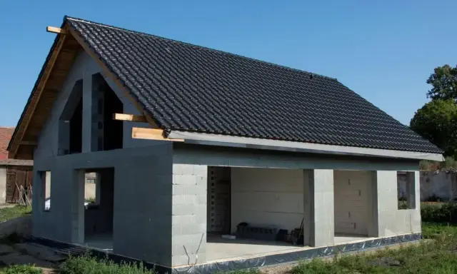 Dom w stanie surowym otwartym, z betonowych pustaków, z dachem pokrytym czarną dachówką. Widać otwory okienne i drzwiowe.