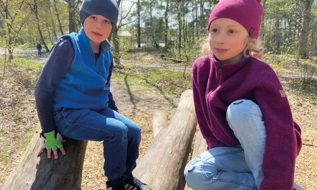 Två barn sitter på stockar i skogen. Båda bär en mjuk merinoull mössa barn, perfekt för utomhuslek.