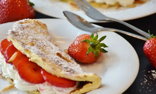 Puszysty omlet z białek, nadziany kremem i truskawkami, posypany cukrem pudrem. Obok leżą świeże owoce i sztućce.