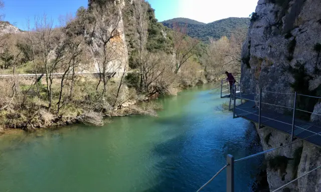 Persona camina por pasarela metálica junto a río turquesa, explorando las rutas de Burgos. Paisaje rocoso y vegetación de ribera.