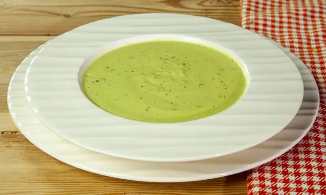 Una ciotola di cremosa crema di zucchine, condita con pepe nero, servita su piatti bianchi su un tavolo di legno.