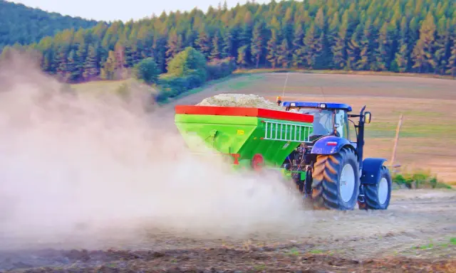 Traktor rozsiewa wapno do ziemi, tworząc chmurę pyłu na polu. W tle las i wzgórza.