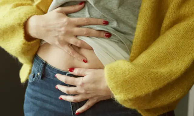 En kvinna med röda naglar håller om sin mage, klädd i en gul kofta och jeans. Hon känner sig gravid trots mens.