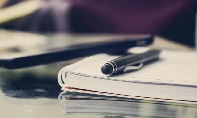 Czarny długopis spoczywa na otwartym notatniku. Obok leży laptop, gotowy do pracy. FGŚP.
