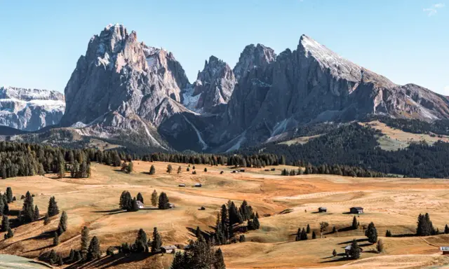 Un paesaggio mozzafiato delle Dolomiti più belle, con cime imponenti, prati dorati e boschi verdi. Piccole baite sparse creano un'atmosfera idilliaca.