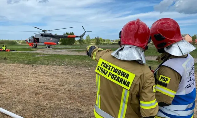 Strażacy w akcji. W tle lądujący śmigłowiec, który wspiera pracę w straży pożarnej.