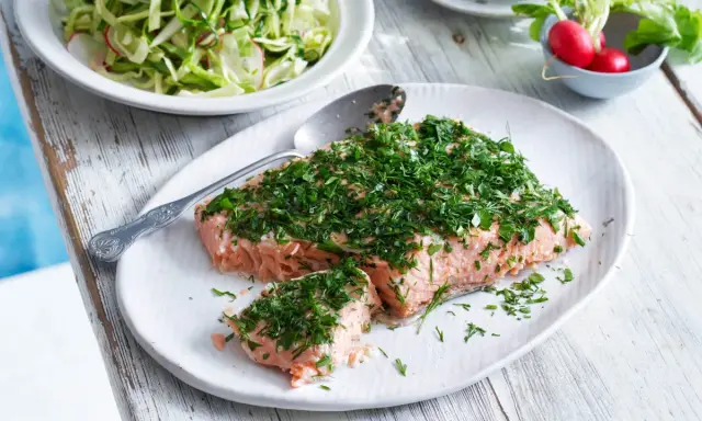 En saftig bit confiterad lax, generöst toppad med färska örter, serveras på en vit tallrik. Bredvid syns en fräsch sallad och några röda rädisor.