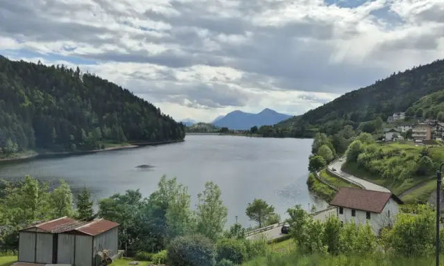 Panoramica del lago di Stramentizzo circondato da montagne verdi e un villaggio pittoresco. Nuvole minacciose nel cielo.