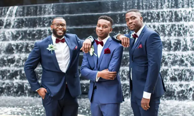 Three groomsmen in stylish navy blue suit combinations, featuring burgundy bow ties and pocket squares, pose in front of a waterfall.