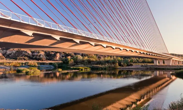 El puente de Castilla-La Mancha se refleja en el río, con sus cables rojos y el cielo azul.
