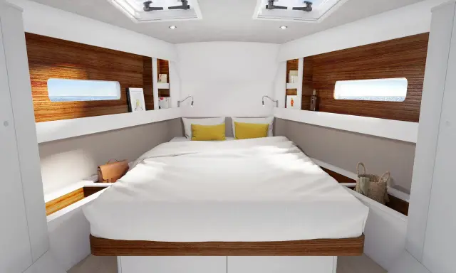 L'intérieur d'un voilier 8m, avec un grand lit blanc, des coussins jaunes, des hublots donnant sur la mer et des étagères en bois.