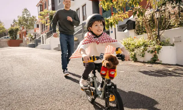 Un bambino con un mantello da supereroe prova ad andare in bicicletta, mentre suo padre corre dietro per aiutarlo. Mio figlio non riesce a pedalare, ma si diverte un mondo!