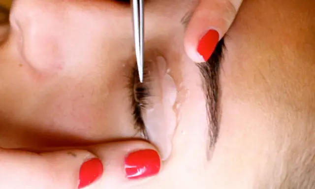 Application d'un produit pour recourber les cils. Une main aux ongles rouges tient délicatement la paupière pendant que l'outil travaille.