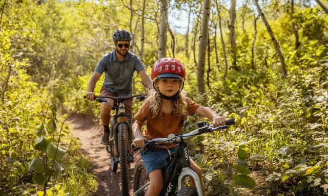 En flicka och en man cyklar på en stig i en lummig skog. Solen lyser genom träden och skapar ett vackert ljusspel.