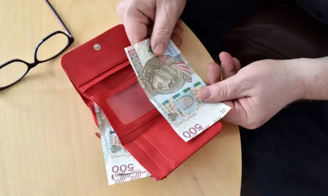 Ręce wkładają banknot 500 zł do czerwonego portfela. Obok okulary. Może to być pomocne przy sprawdzaniu 14 emerytury tabeli netto.
