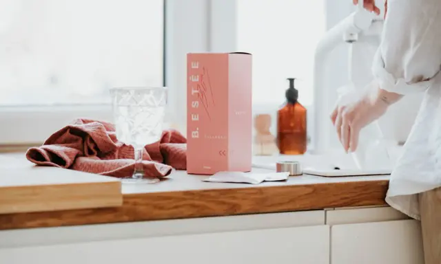 Różowe pudełko z kolagenem B. ESTÉE, szklanka wody i ręcznik. To estetyczne zdjecia produktowe.