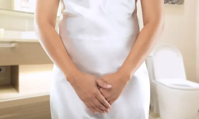 Kobieta w białej sukience w łazience, z dłońmi skrzyżowanymi na brzuchu. W tle toaleta. Szuka domowych sposobów na nietrzymanie moczu.