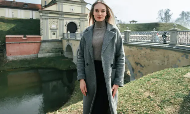 Kobieta w szarym płaszczu i golfie, idealna stylizacja na 14 stopni. W tle zamek i most.