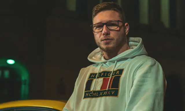 Kubańczyk cały ja utwory. Młody mężczyzna w okularach i bluzie z kapturem Tommy Hilfiger, nocą, obok żółtego samochodu.