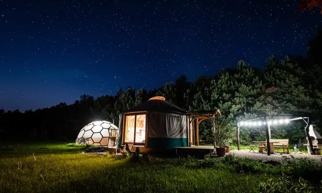 Nocne niebo nad Jurą Krakowsko-Częstochowską. Dwa noclegowe domki, jurt i kopuła, oświetlone ciepłym światłem, otoczone zielenią.