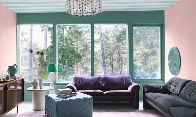 En färgstark salong med 30-tals inredning. Mörklila soffa, grå soffa, turkos pall och rosa matta. Fönster med utsikt över skog.