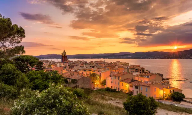 Sonnenuntergang über St. Tropez. Die Sonne versinkt im Meer, beleuchtet die Stadt und den Kirchturm in warmen Farben.