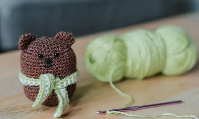 Os **sonajeros de crochet patrones gratis** son adorables. Un osito marrón con bufanda verde y blanca, junto a ovillos de lana y una aguja de crochet.