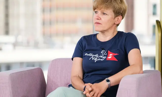 Monika Witkowska w granatowej koszulce z napisem "Odbudujmy Nepal" siedzi w różowym fotelu, patrząc w bok.